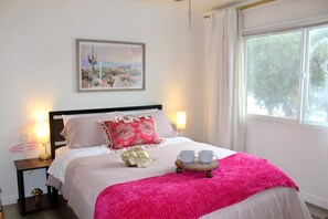 Ein ordentlich bezogenes Bett mit einer rosa Decke, einem Zierkissen und einem Tablett mit zwei Tassen. An der Wand hängt ein Bild und ein Fenster mit Vorhängen.