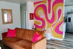 Una sala de estar con un sofá marrón, un muro abstracto y colorido, y una cocina con armarios y un refrigerador.