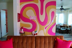Ein Wohnzimmer mit einem farbenfrohen Wandgemälde, einem braunen Sofa mit roten Kissen und einem kleinen Tisch mit Dekoration.
