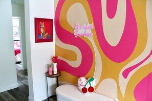 Ein Zimmer mit einem lebendigen Wandgemälde, einem kleinen Tisch mit einer Vase und Blumen sowie einem herzförmigen Kissen mit Namensschild.
