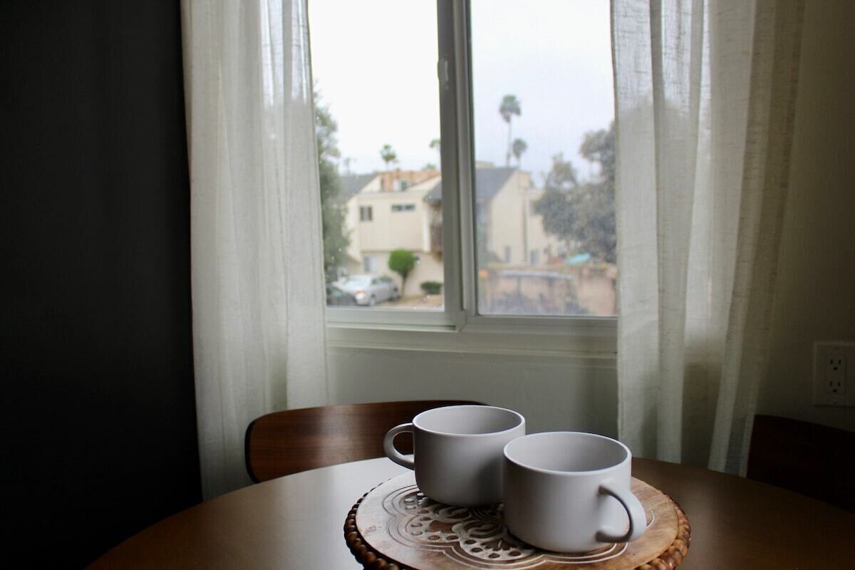 A table with two white mugs on a wooden coaster by a window with white curtains.