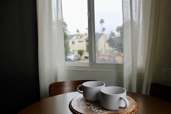 Una mesa con dos tazas blancas sobre un posavasos de madera, cerca de una ventana con cortinas blancas.