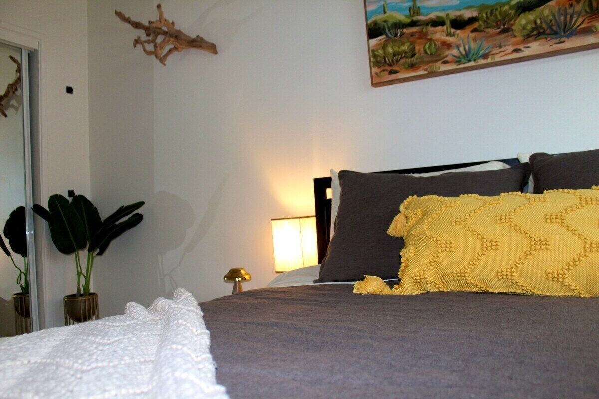 A bed with a grey blanket and a yellow pillow, a bedside lamp, a plant, and a wall-mounted artwork.
