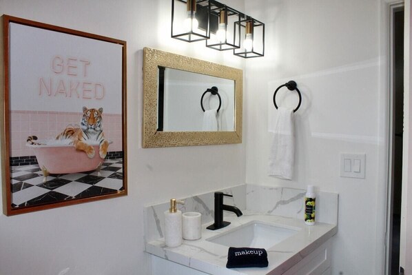 A bathroom with a sink, mirror, and towel rings. There is a poster on the wall and a bottle of lotion on the sink.