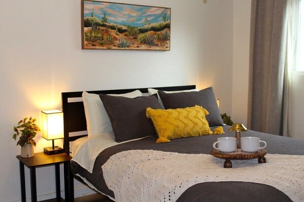 Una cama bien tendida con una manta blanca, una almohada gris y amarilla, y una mesita de noche con una lámpara y una planta.