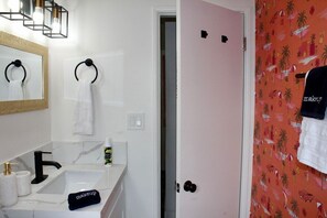 Ein Badezimmer mit einem weißen Waschbecken, schwarzen Handtuchhaltern, einem Spiegel mit goldener Rahmung und einer Tür zu einem Raum mit tropischem Wandpapier.