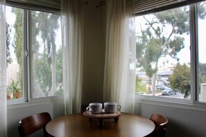 Ein Eckzimmer mit Tisch und zwei Stühlen, einem Fenster mit Vorhängen und Blick auf Bäume und Gebäude.