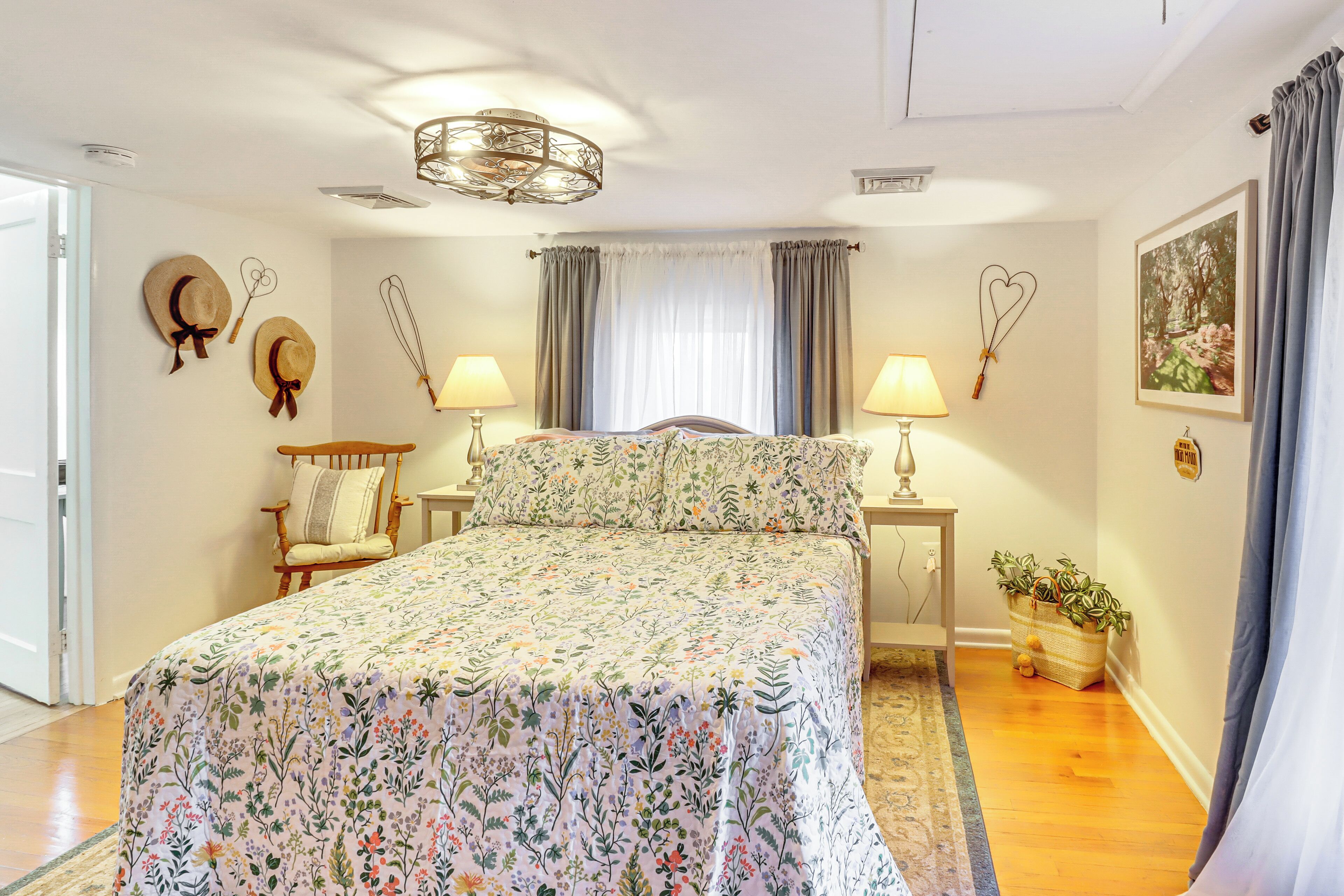 A bedroom with a floral bedspread, a wooden chair, two bedside tables with lamps, a chandelier, and a window with curtains.