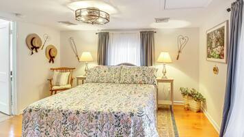 A bedroom with a floral bedspread, a wooden chair, two bedside tables with lamps, a chandelier, and a window with curtains.