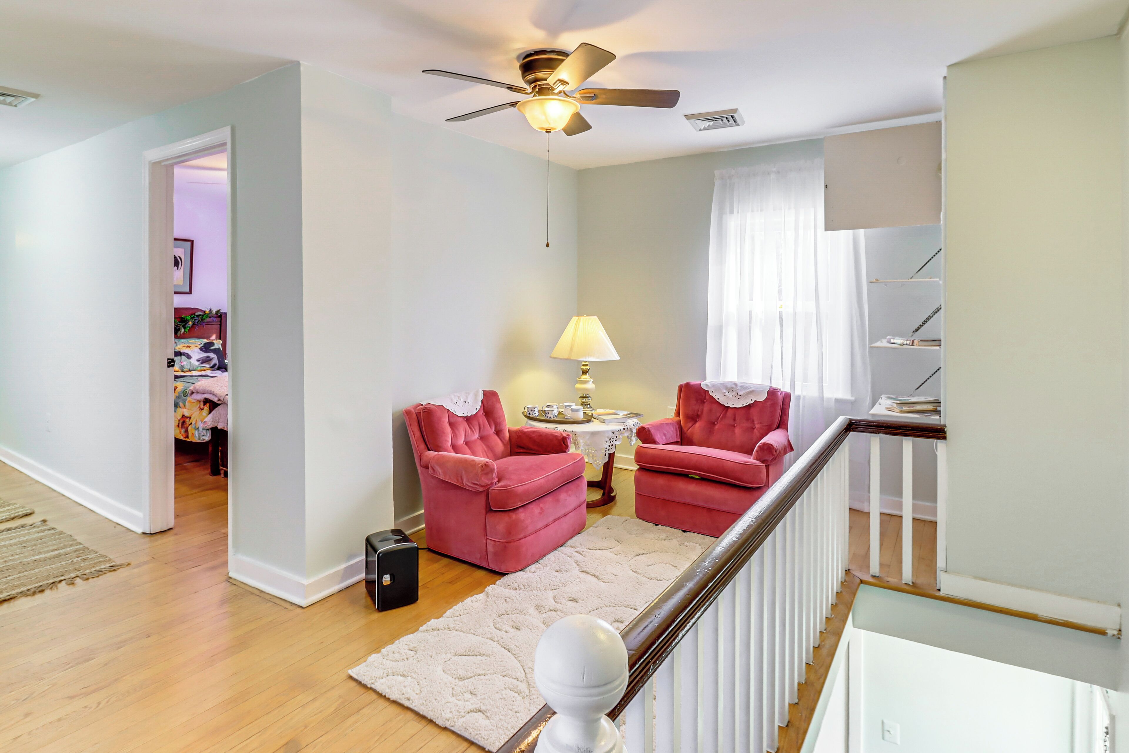 A spacious living room with a ceiling fan, two pink armchairs, a white rug, and a staircase with a white railing.