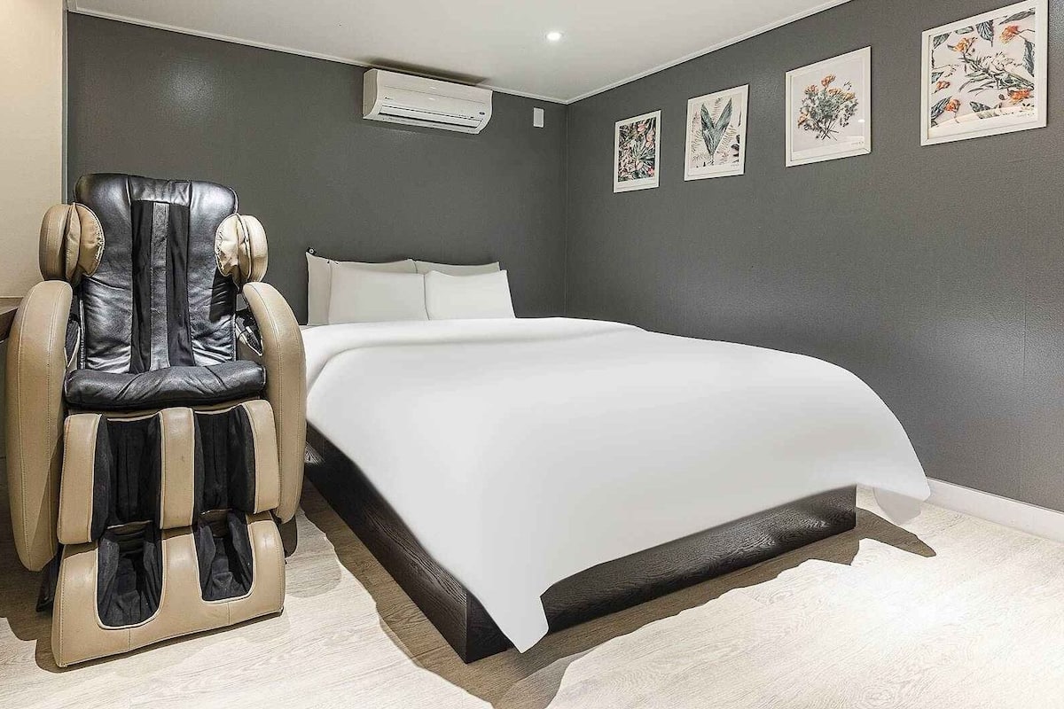 Un dormitorio con sillón de masaje, cama, aire acondicionado de pared y láminas botánicas enmarcadas.