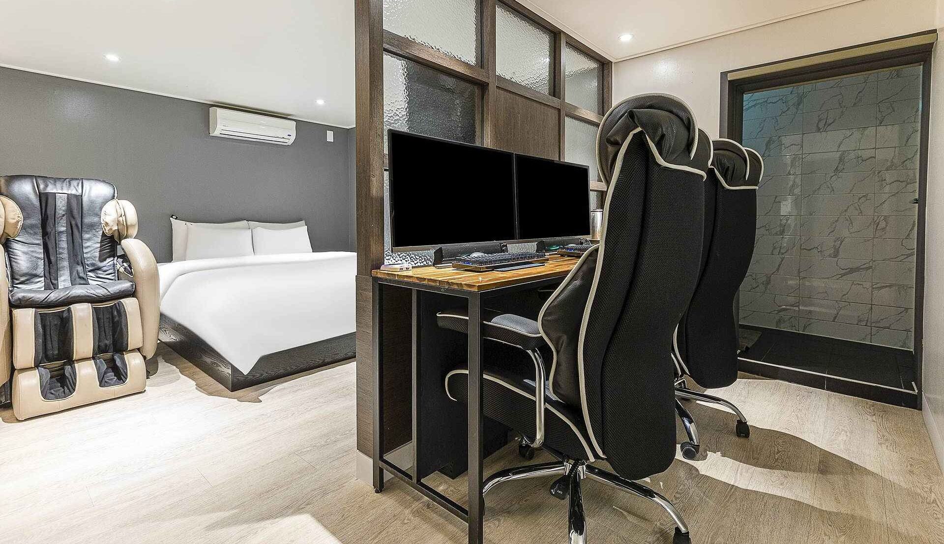 A modern hotel room with a bed, a desk with a computer, a massage chair, and a large window with a stone wall view.