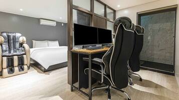 A modern hotel room with a bed, a desk with a computer, a massage chair, and a large window with a stone wall view.