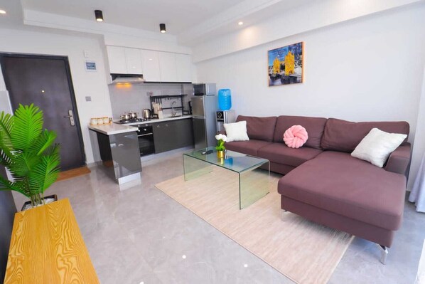 Een moderne woonkamer met een glazen salontafel, een bruine bank en een keuken op de achtergrond.