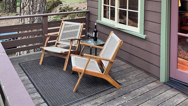 Una terraza de madera con dos sillas de mimbre y una mesa, una botella de vino y un farol encendido.