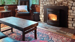 Un acogedor interior de cabaña con una chimenea de piedra, una zona de estar confortable con un banco y una silla, y un ventanal con vistas al bosque circundante.