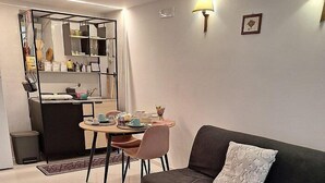 A modern living room with a dark grey sofa, a small round table, and a kitchen area with a built-in fridge and shelves.