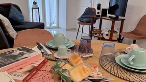 A dining area with a table set for breakfast, including bread, pastries, and coffee. There are chairs around the table, and a TV is visible in the background.