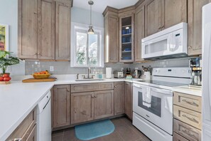 Una cocina con armarios de madera, encimera blanca, microondas empotrado, horno y ventana con vista a árboles.