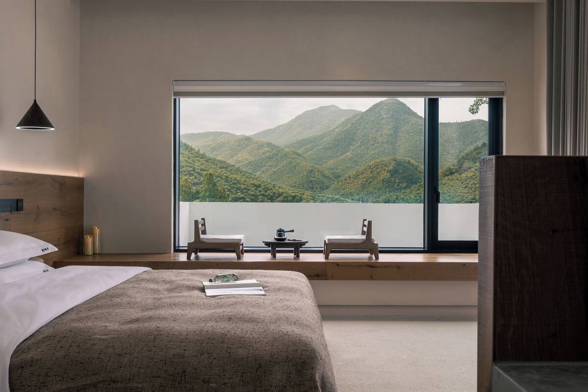 A bedroom with a large window, a bed, two chairs, a small table, and a view of a mountainous landscape.