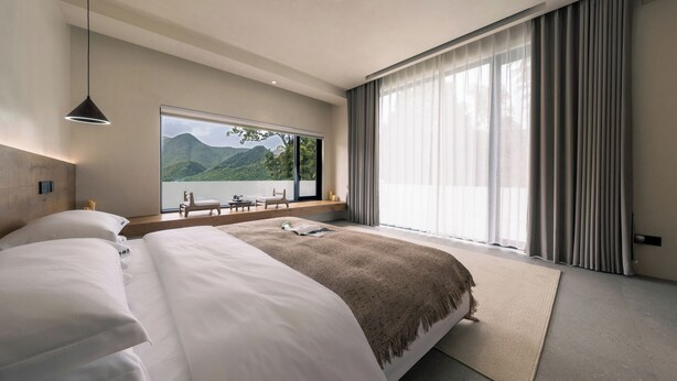 Een moderne hotelkamer met een groot bed, uitzicht op bergen door een raam, en een balkon met een tafel en stoelen.