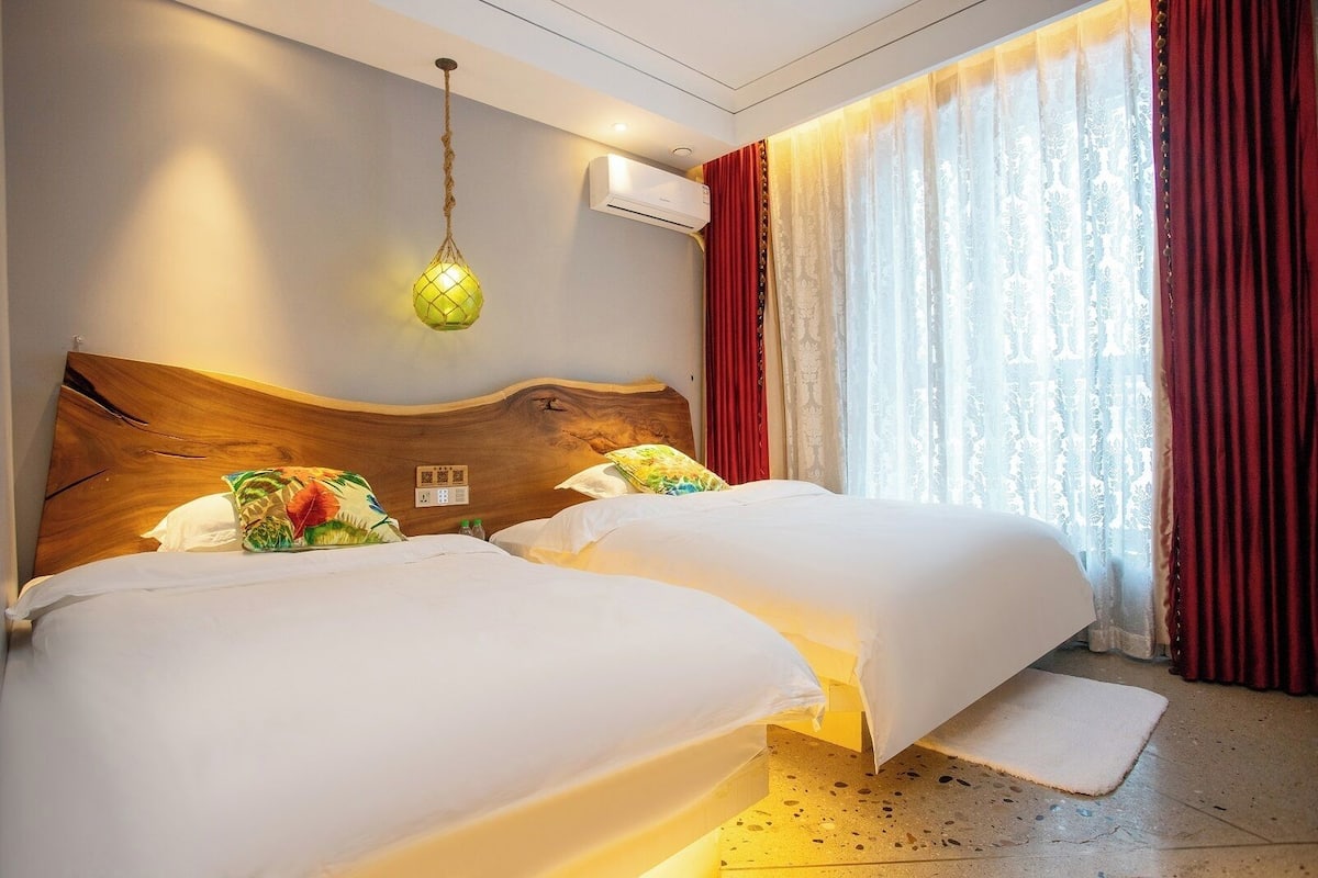 Habitación de hotel con dos camas, cabecera de madera, lámpara colgante, ventana con cortinas rojas y equipo de aire acondicionado.