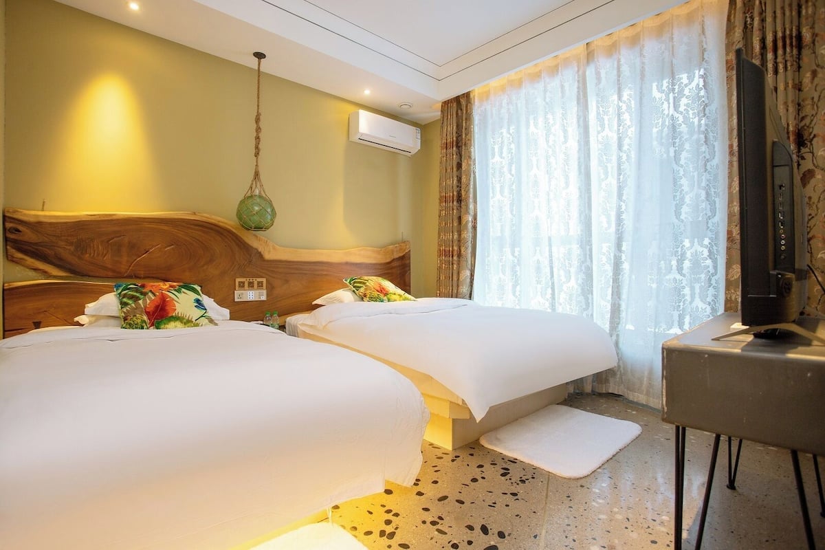 Habitación de hotel con dos camas, cabecera de madera, lámpara colgante, unidad de aire acondicionado, televisor y ventana con cortinas.