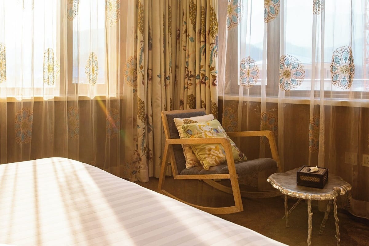 Un dormitorio con una cama, un sillón mecedor, una mesita y cortinas con motivos florales.