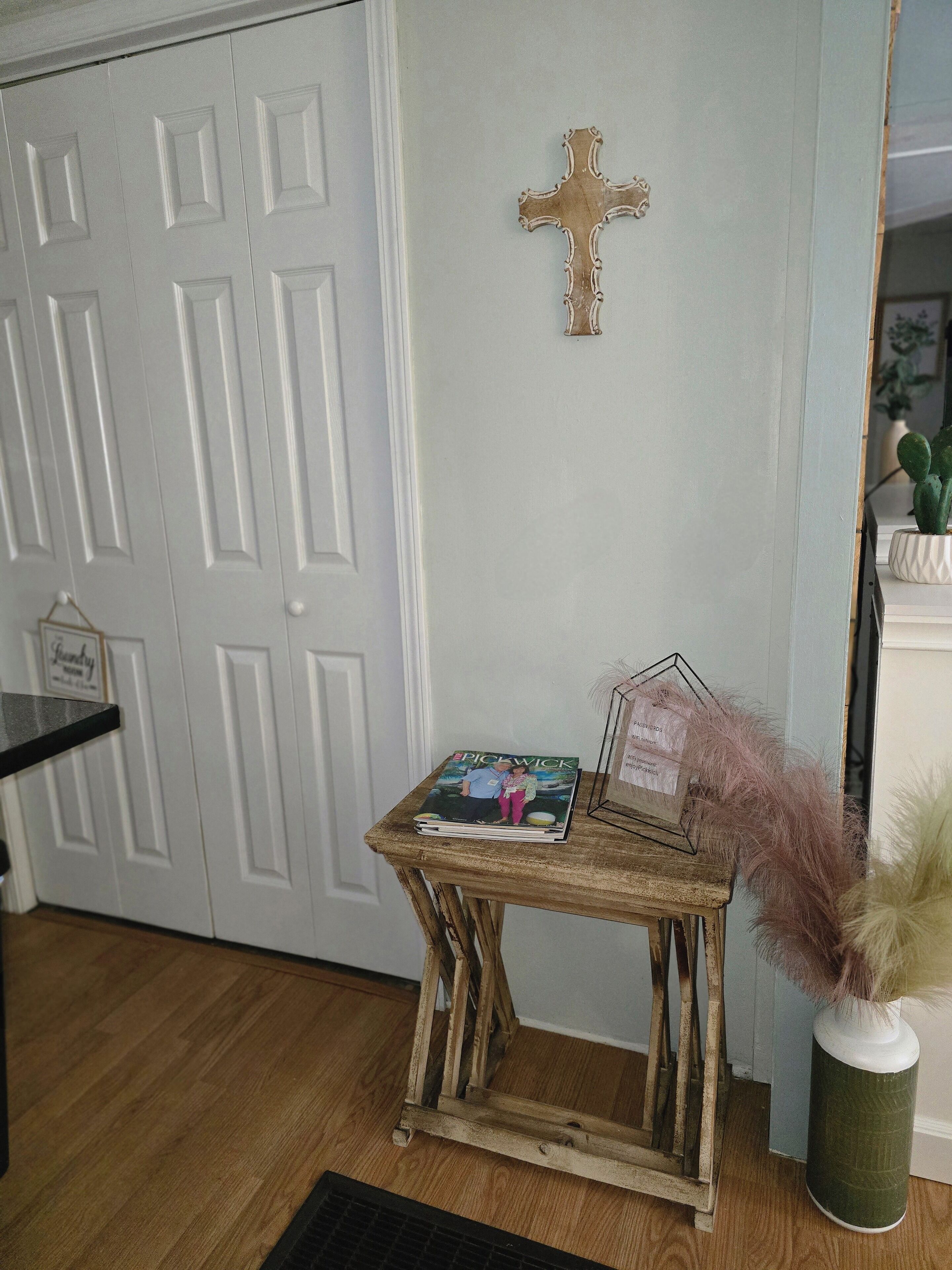 A wooden side table with a magazine, a decorative cross on the wall, and a cactus in a vase.