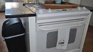 Pieni, käytännöllinen keittiö, jossa on valkoinen liesi, musta roskakori ja puinen leikkuulauta työtasolla.