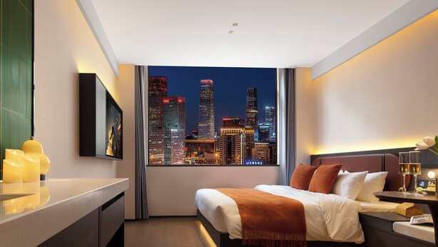 A modern hotel room with a cityscape view, a bed with a white and brown blanket, a bedside table with a glass of wine, and a nightstand with a lamp. - Merica Hotel -Chengdu Chunxi Road (Chengdu)