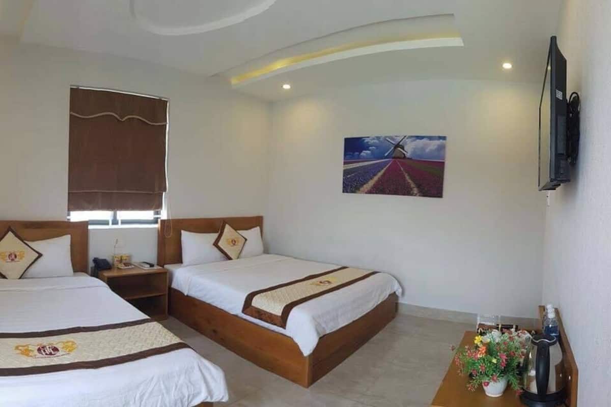 Habitación de hotel con dos camas, una mesita con un florero, un cuadro en la pared y un televisor montado.