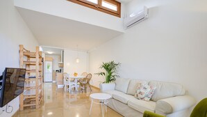 A modern living room with a white sofa, a green armchair, a small white table, and a wooden bookshelf. There is a dining area with a table and chairs in the background. - Comtat Muro 2 by Rentallorca (Muro)