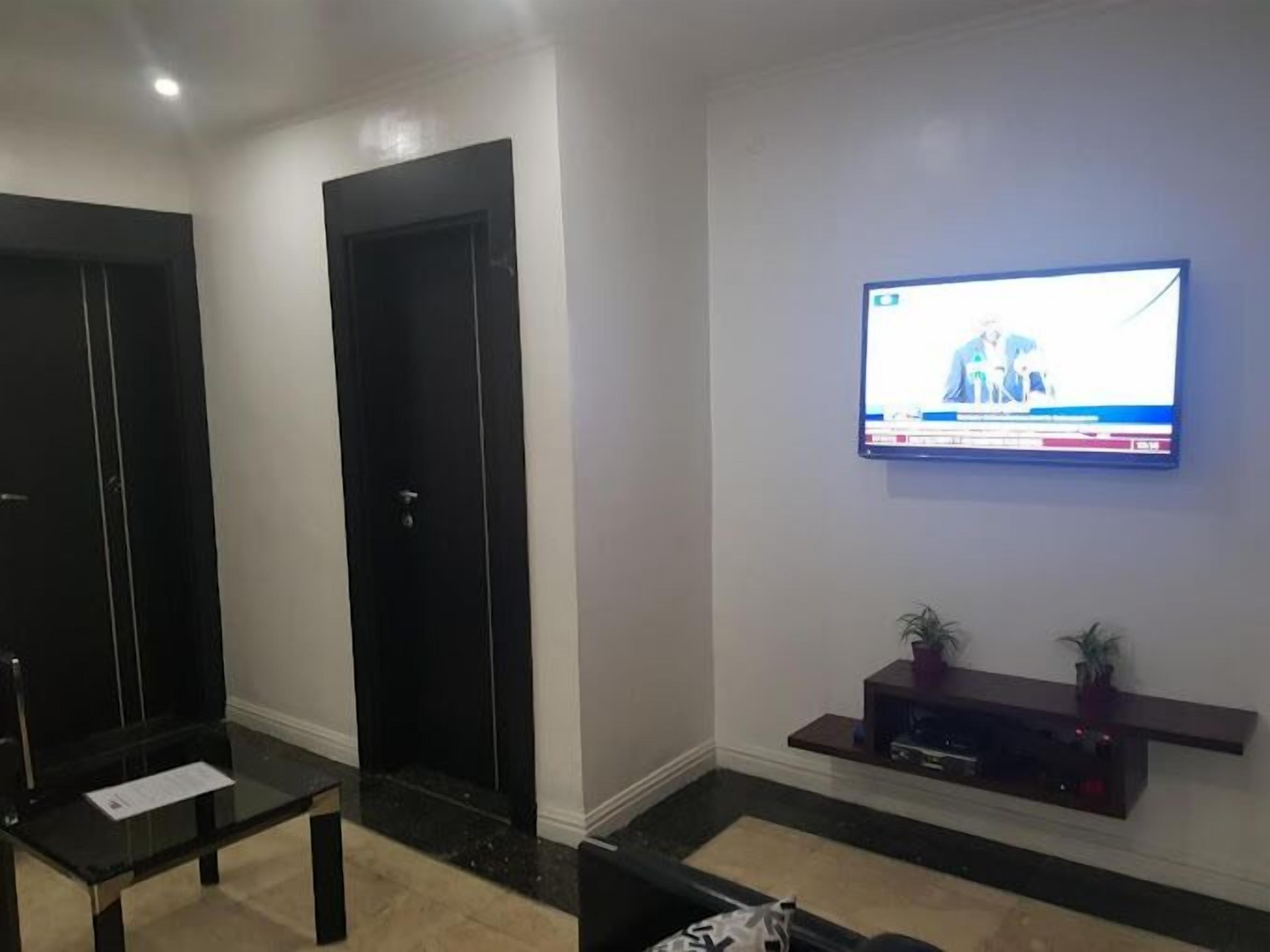 Een moderne woonkamer met een flatscreen televisie aan de muur, een donker houten deur en een glazen salontafel.