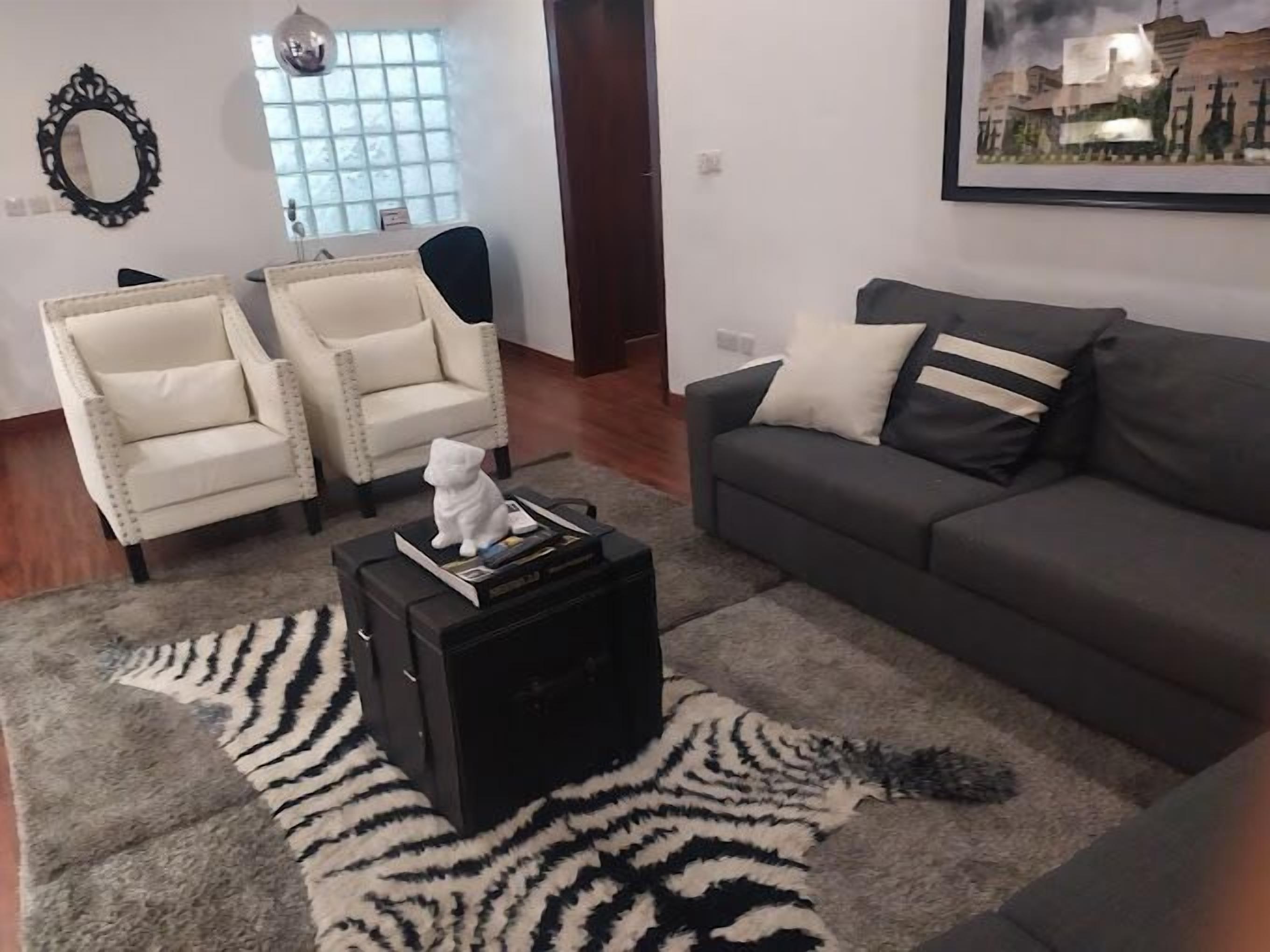 A living room with a grey sofa, two white armchairs, a black coffee table with a white cat figurine, a zebra-patterned rug, and a framed cityscape painting.
