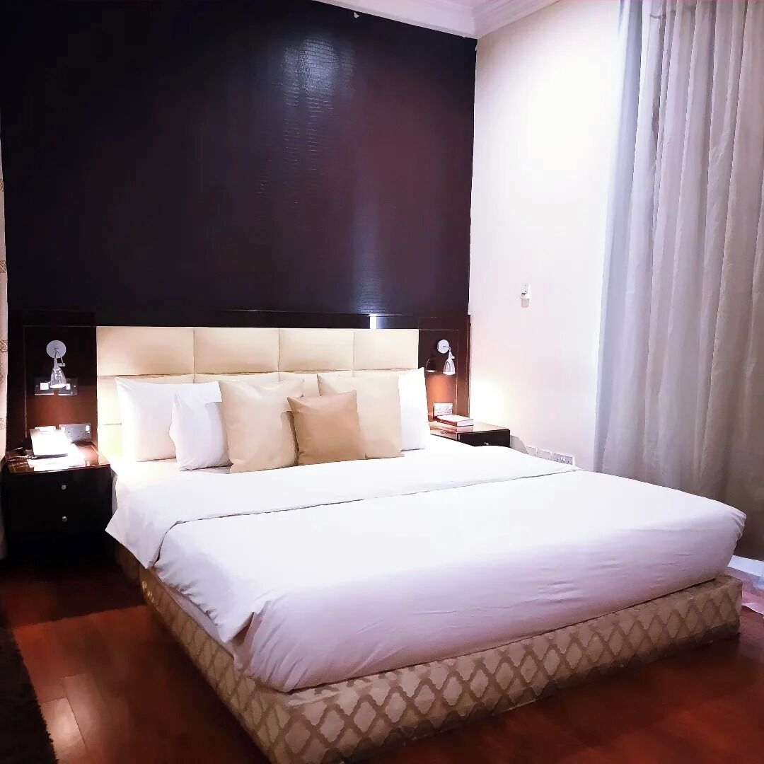 Una habitación de hotel con una cama grande, un cabecero oscuro y una mesita de noche con lámpara.