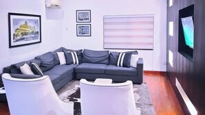 A modern living room with a sectional sofa, two white armchairs, a flat-screen TV mounted on the wall, and framed pictures on the walls. - NiCotel Apartments (Abuja)