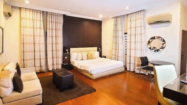 A modern hotel room with a large bed, a sofa, a small table, and a chair. - NiCotel Apartments (Abuja)