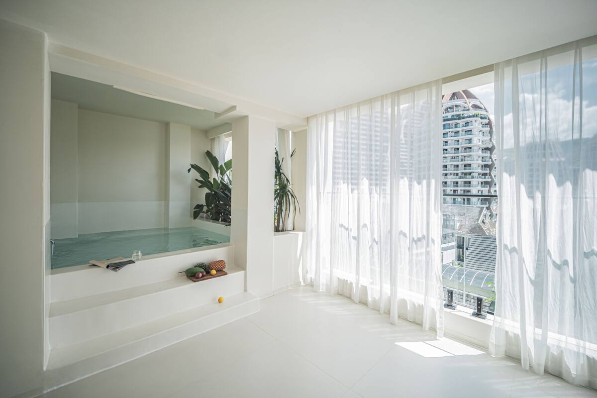 Une salle de bain moderne avec une douche en verre, un grand miroir et une vue sur un immeuble de grande hauteur à travers des rideaux transparents.