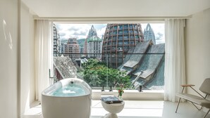 Ein modernes Badezimmer mit einer großzügigen Badewanne, einem kleinen Tisch mit einer Vase und Blick auf eine Stadtlandschaft durch große Fenster.