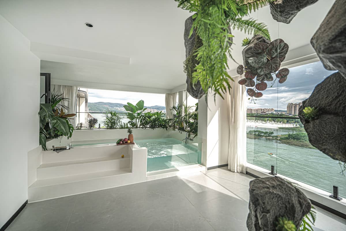 Un intérieur moderne avec un jacuzzi entouré de parois de verre, de grandes fenêtres offrant une vue sur un plan d’eau et des bâtiments éloignés, ainsi que des plantes d’intérieur.