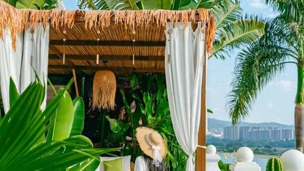 Eine Frau in einem weißen Kleid und Strohhut steht an einem hölzernen Gazebo mit weißen Vorhängen, umgeben von üppigen tropischen Pflanzen und mit Blick auf eine Stadtlandschaft.