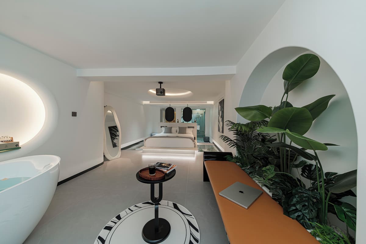 Une chambre d’hôtel moderne avec un grand lit, une table ronde avec un ordinateur portable, une baignoire et un miroir fixé au mur.