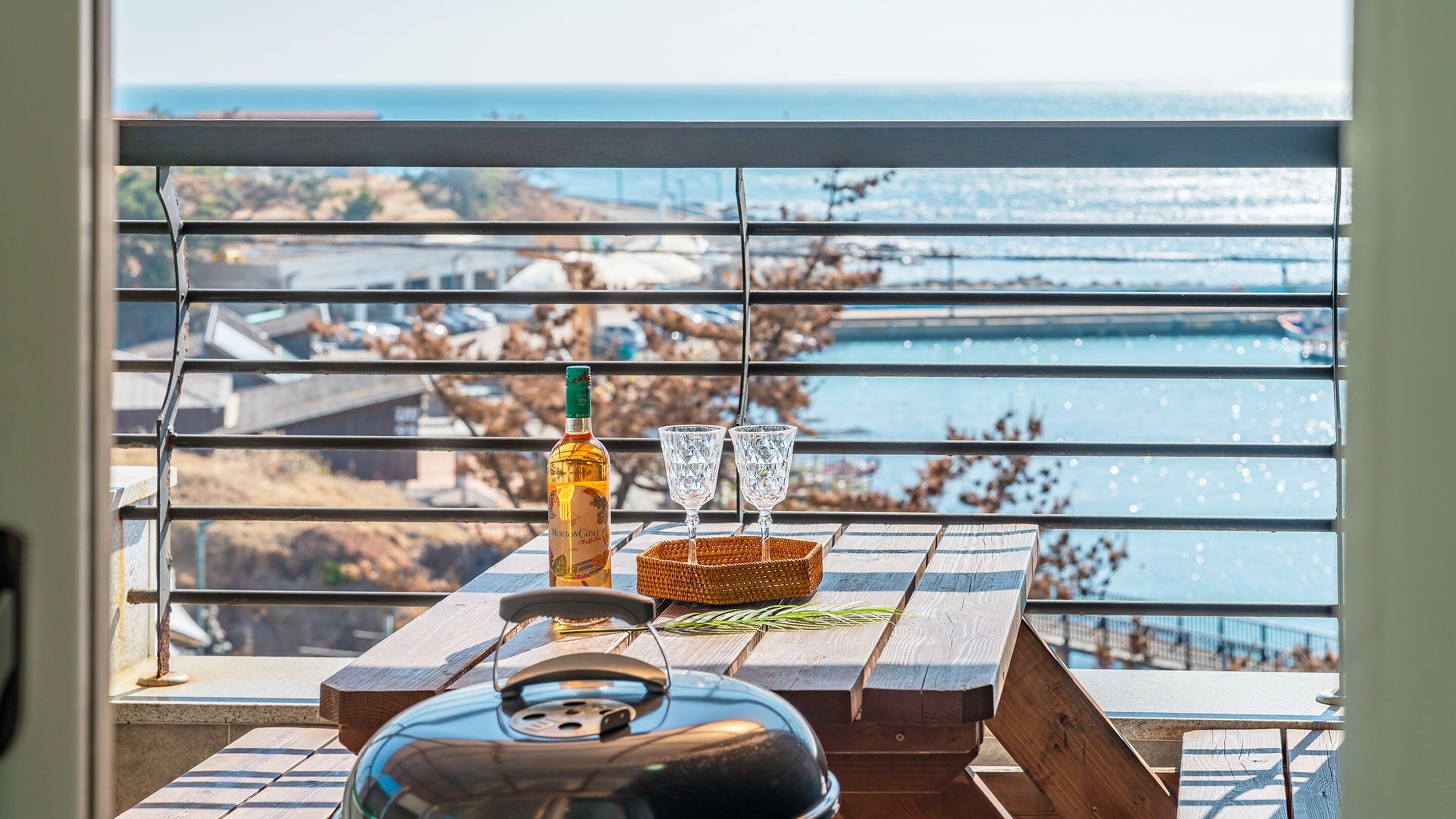 A balcony with a view of the sea, a bottle of liquor, two glasses, and a kettle on a wooden table.