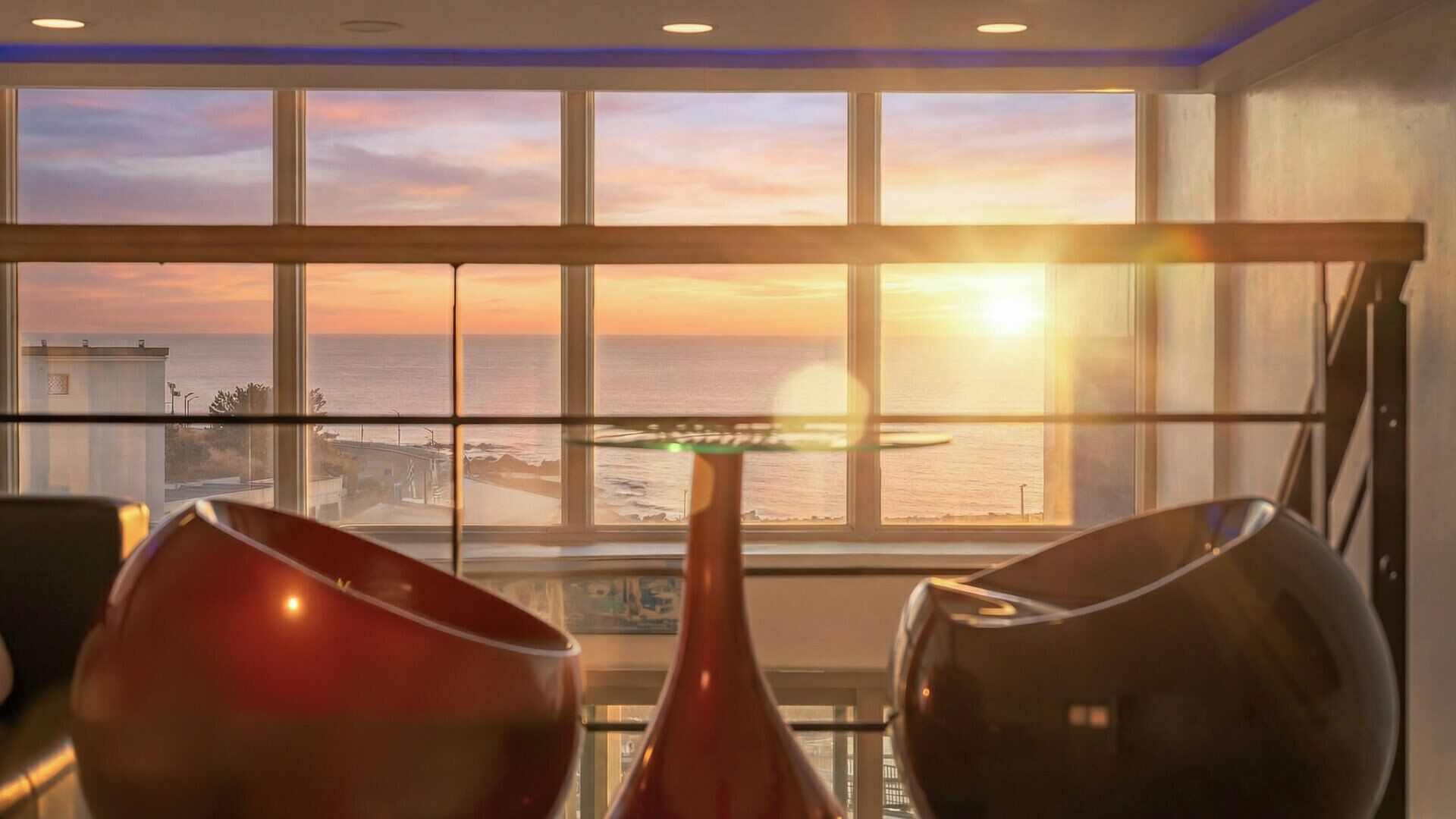 A room with a large window overlooking the ocean at sunset, a glass table, and two chairs.