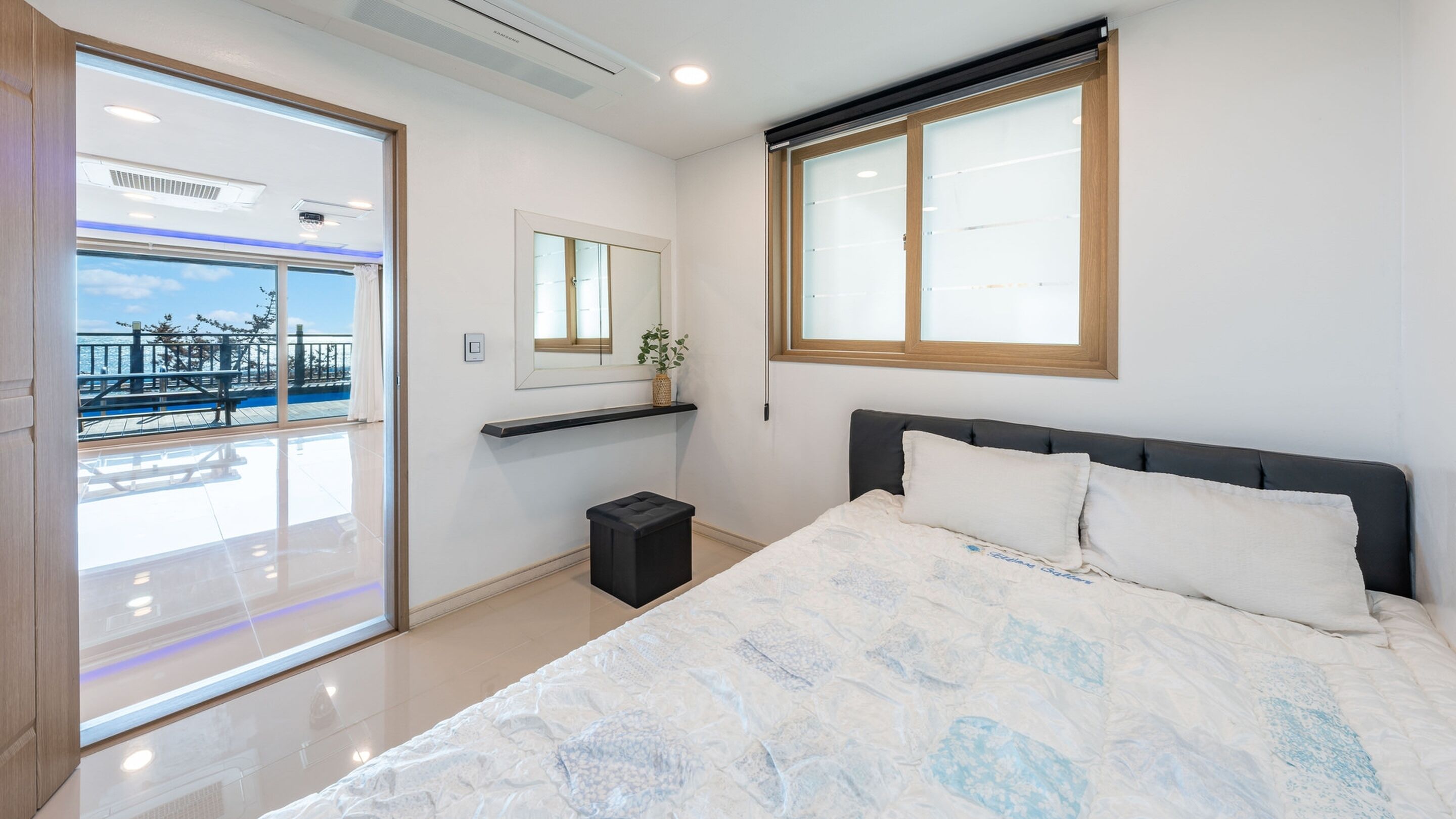 A modern bedroom with a large bed, a small bedside table, a mirror, and a window with a view of a balcony.