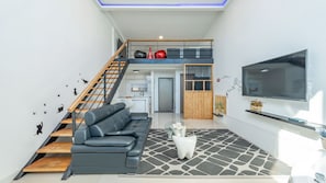 Una sala moderna con un sofá de cuero negro, un pouf blanco y una alfombra estampada. Un televisor está montado en la pared, y hay una escalera que conduce a un altillo.
