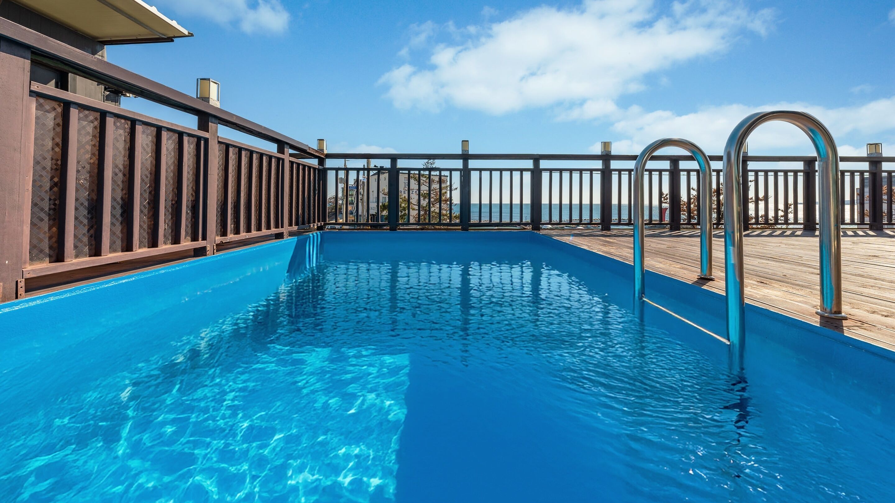 A clear blue swimming pool with a wooden deck and metal railings, overlooking a body of water.