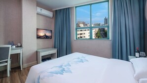 Quarto de hotel com cama, escrivaninha, cadeira, televisão e janela com cortinas.