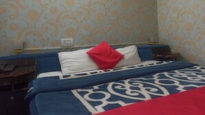 Hotelkamer met bloemenbehang, een bed met een blauw-witte sprei, een rood kussen, en een nachtkastje met een telefoon.